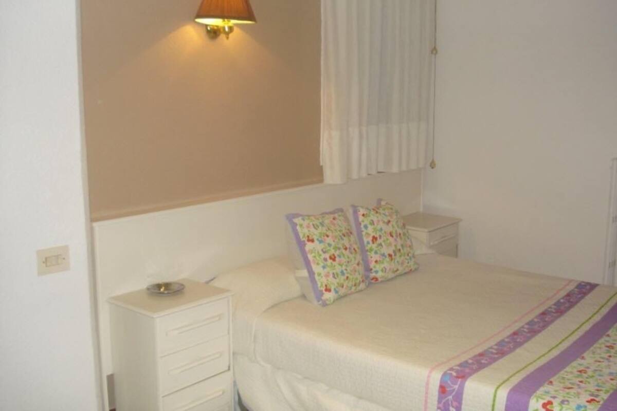 A bedroom with a bed, a bedside table, a lamp, and a window with curtains.