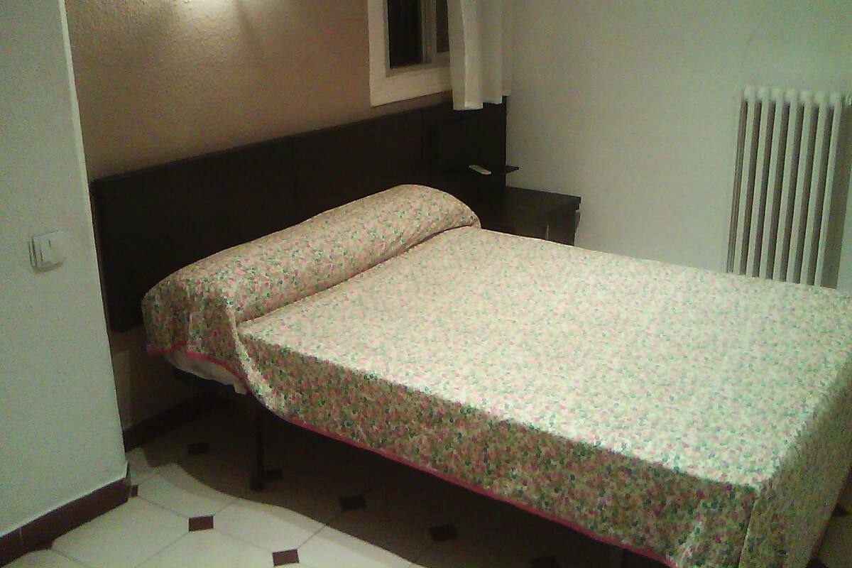 A small, single-bed room with a floral bedspread, a window with curtains, and a radiator.