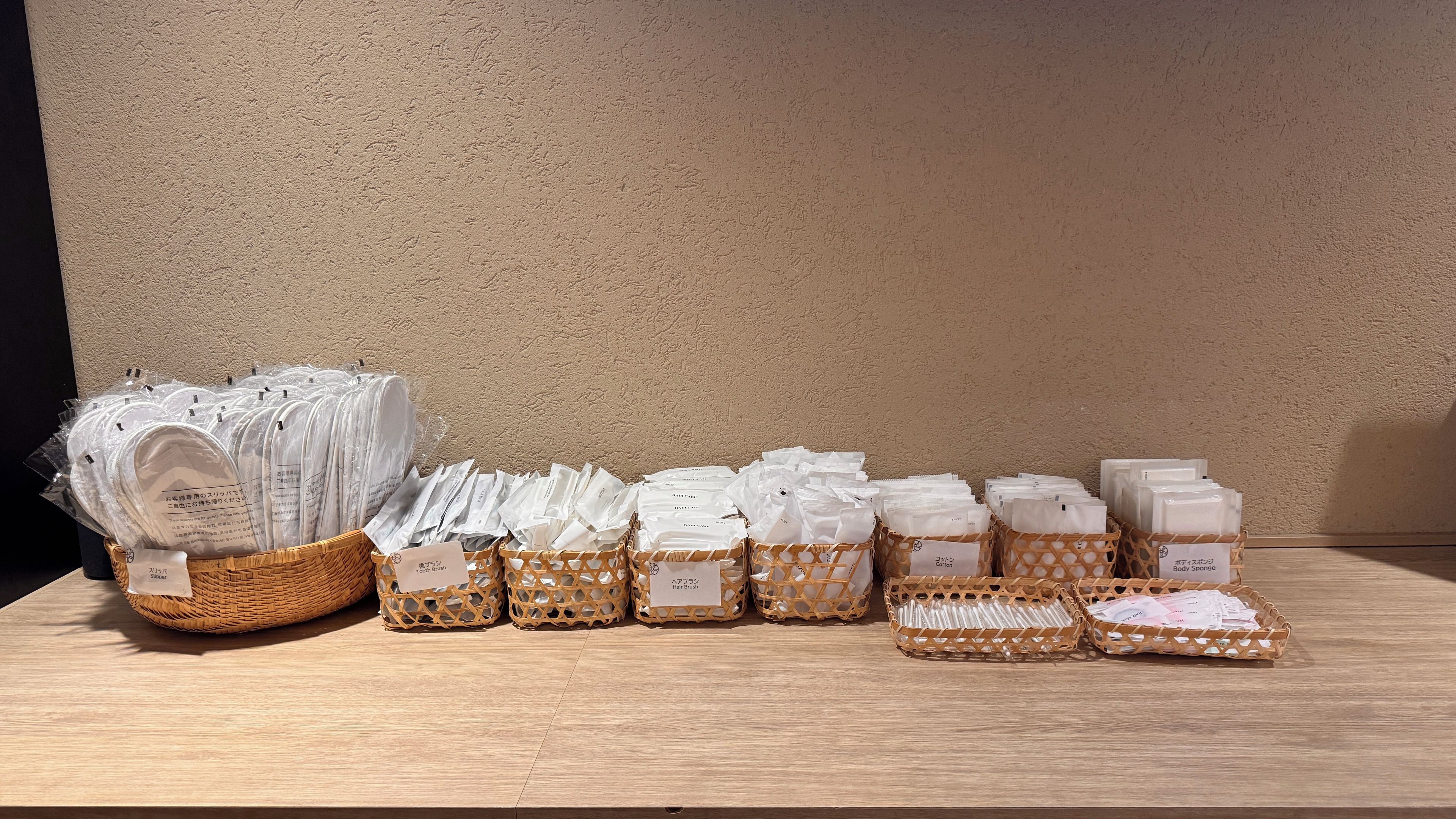 A display of various toiletries in wicker baskets on a wooden table against a textured wall.