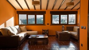 Ein gemütliches Wohnzimmer mit einer Couch, einem kleinen Tisch und Korbstühlen. Große Fenster bieten Blick auf eine Stadt und Hügel.
