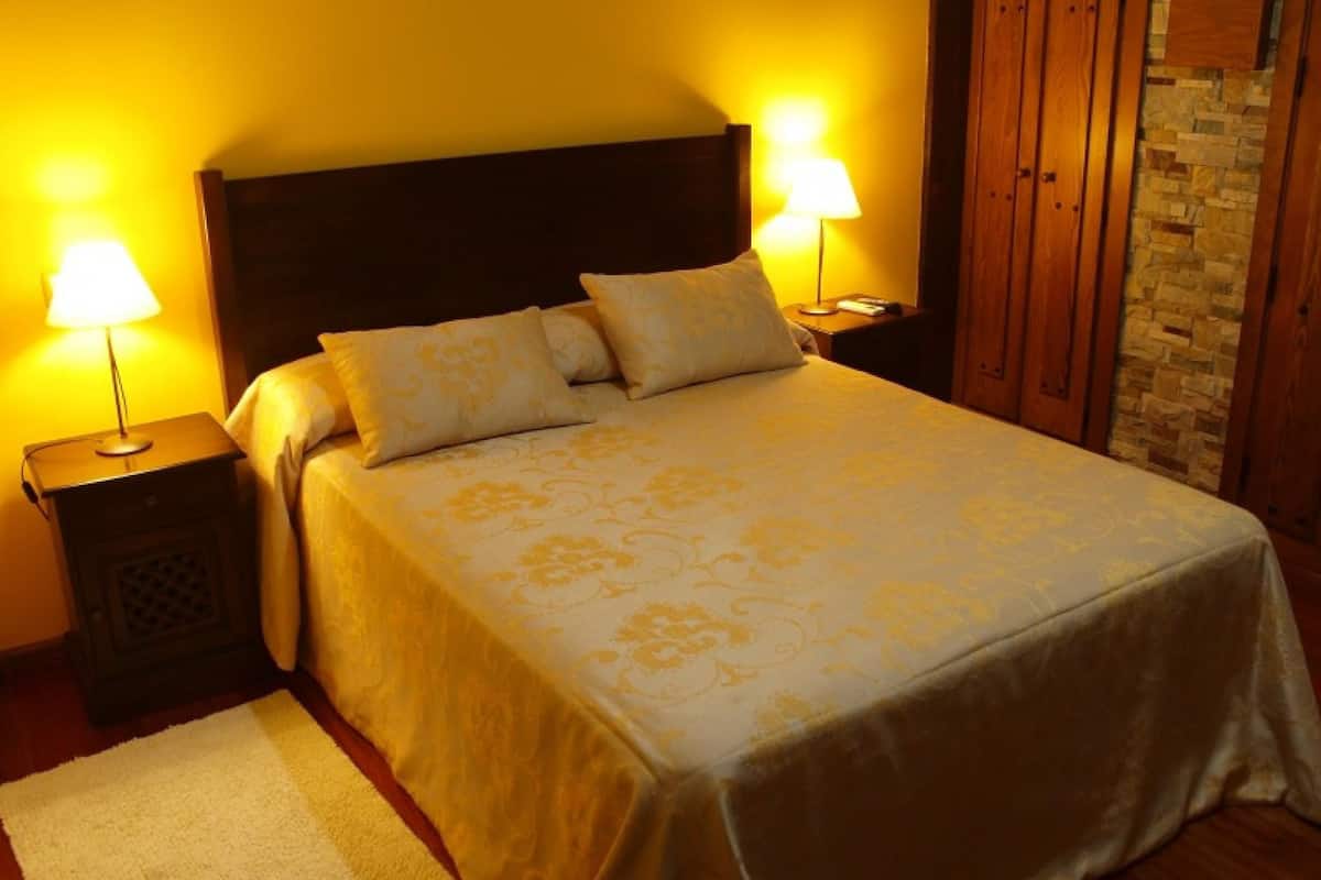 Quarto com cama, duas luminárias de cabeceira, mesa de cabeceira, tapete branco, cabeceira de madeira e parede de pedra.