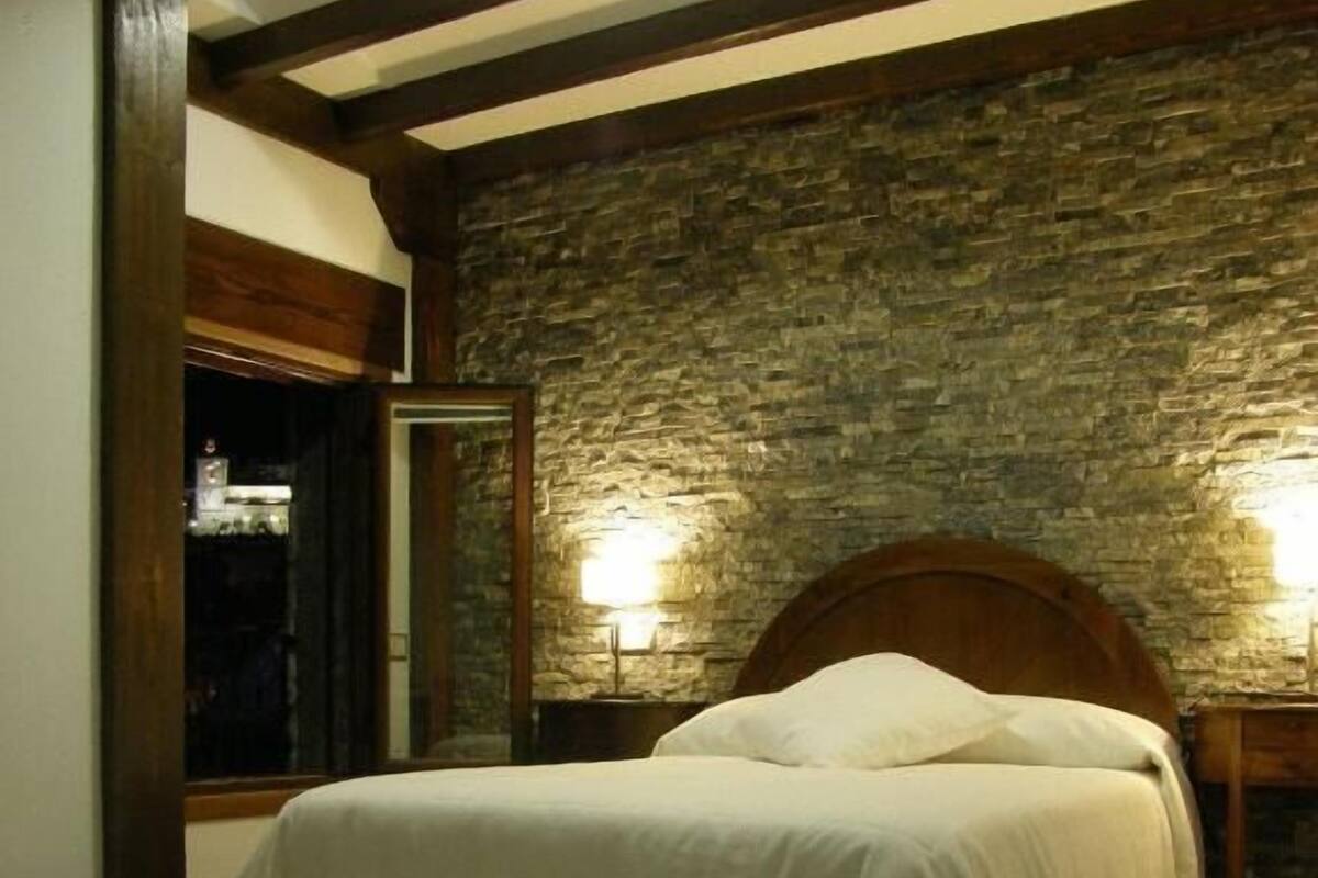 Quarto com parede de pedra, vigas de madeira, cama com roupa de cama branca e duas luminárias de cabeceira.