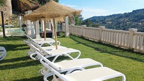 A row of white lounge chairs with a thatched umbrella, a pool, and a view of hilly terrain.