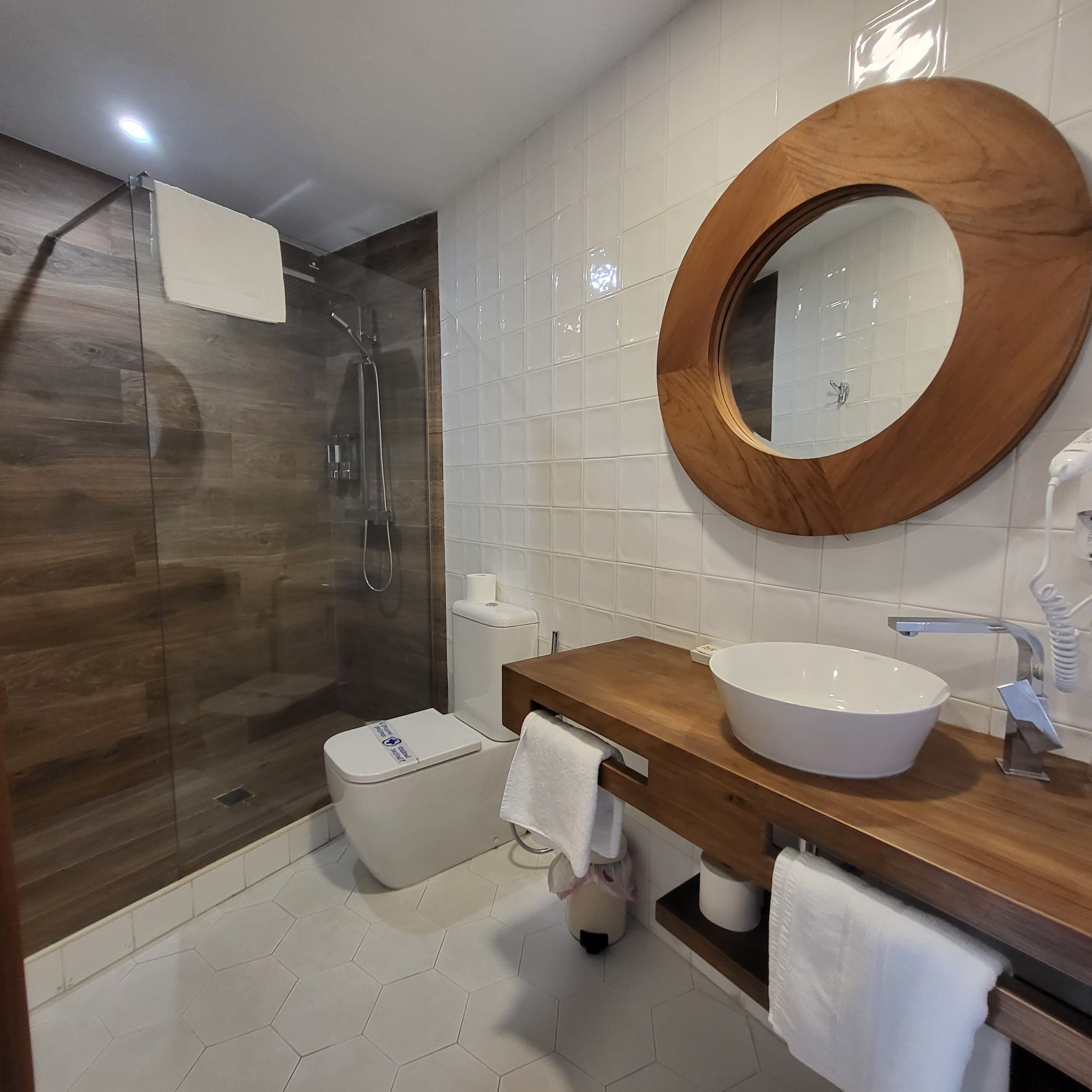 Un baño moderno con ducha de vidrio, tocador de madera con lavamanos grande y espejo redondo.