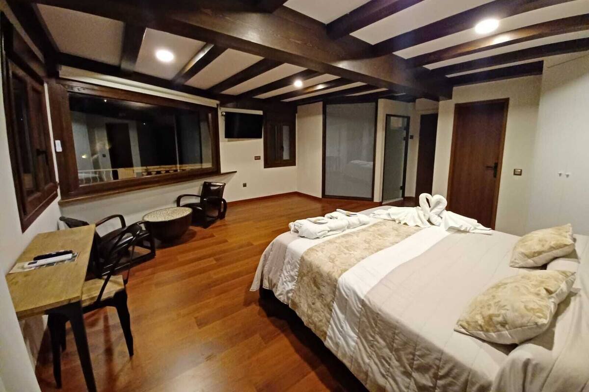 Quarto de hotel com piso de madeira, cama com roupa de cama branca, escrivaninha com cadeira e uma janela grande.