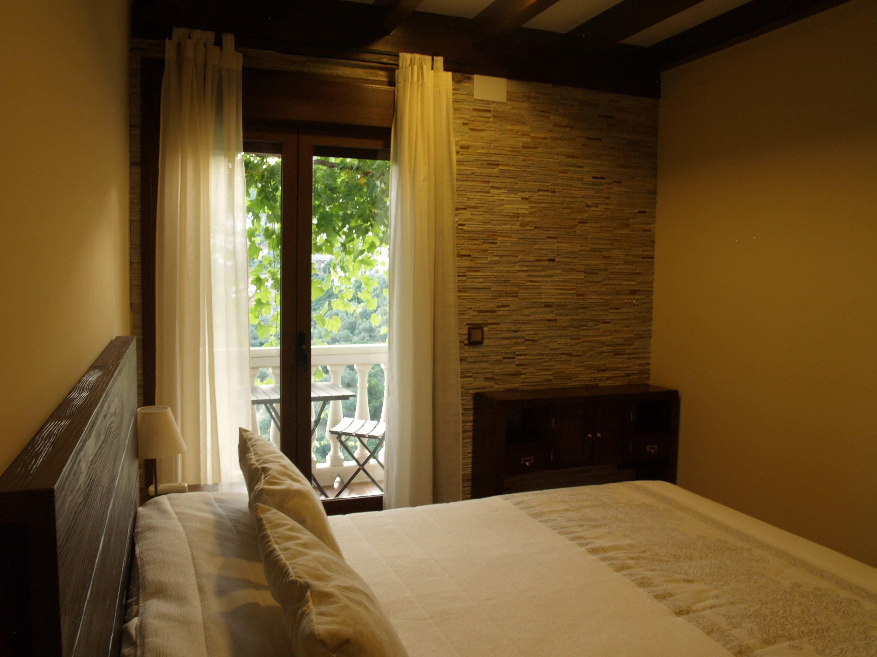 Ein Schlafzimmer mit einem Bett, einem hölzernen Kopfende, einer Steinwand und Blick auf Grün durch ein Fenster.