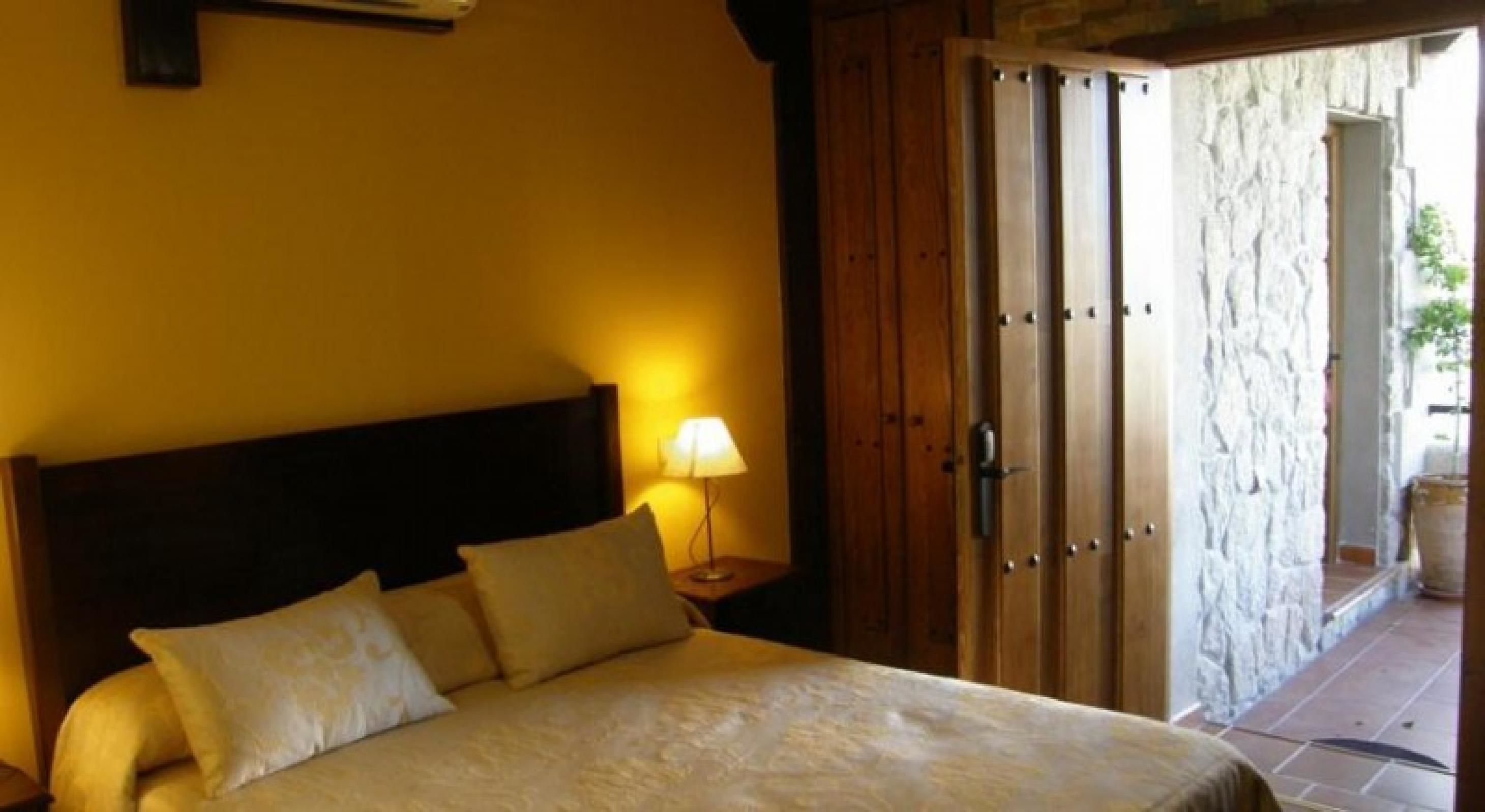 A bedroom with a bed, pillows, a nightstand with a lamp, and a wooden door leading to an outdoor area.