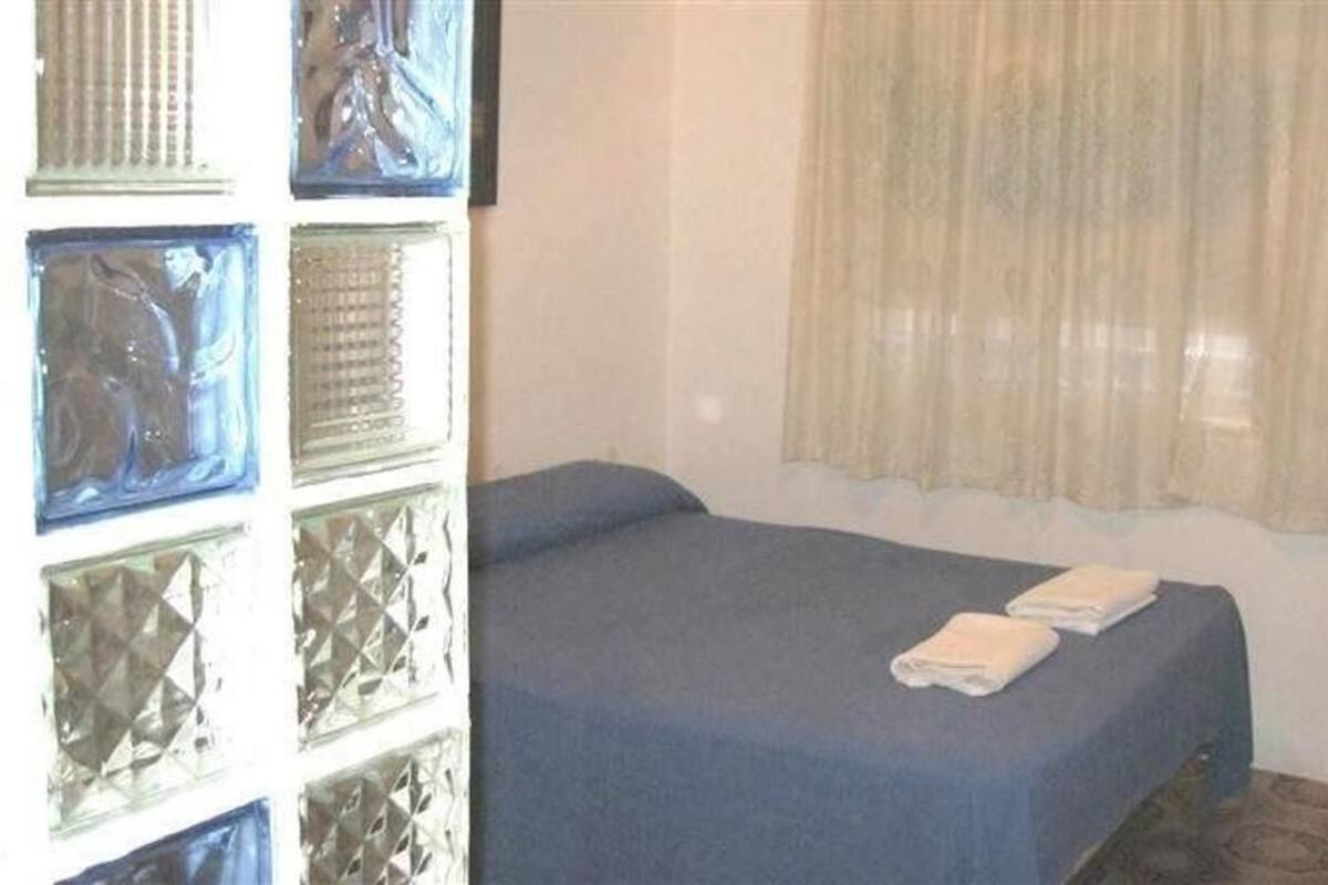 A single bed with a blue bedspread and two folded towels, a window with a curtain, and a wall with decorative glass panels.