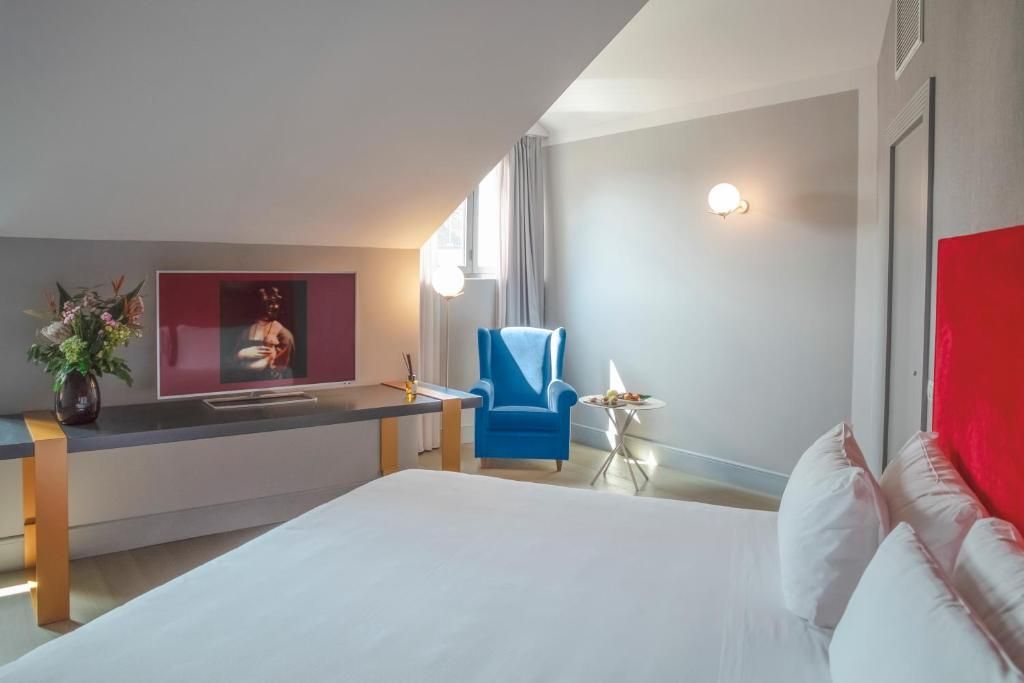A modern hotel room with a bed, a desk, a chair, a small table with a lamp, and a wall-mounted TV.