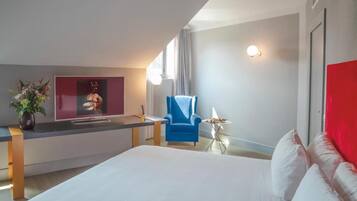 A modern hotel room with a bed, a desk, a chair, a small table with a lamp, and a wall-mounted TV.