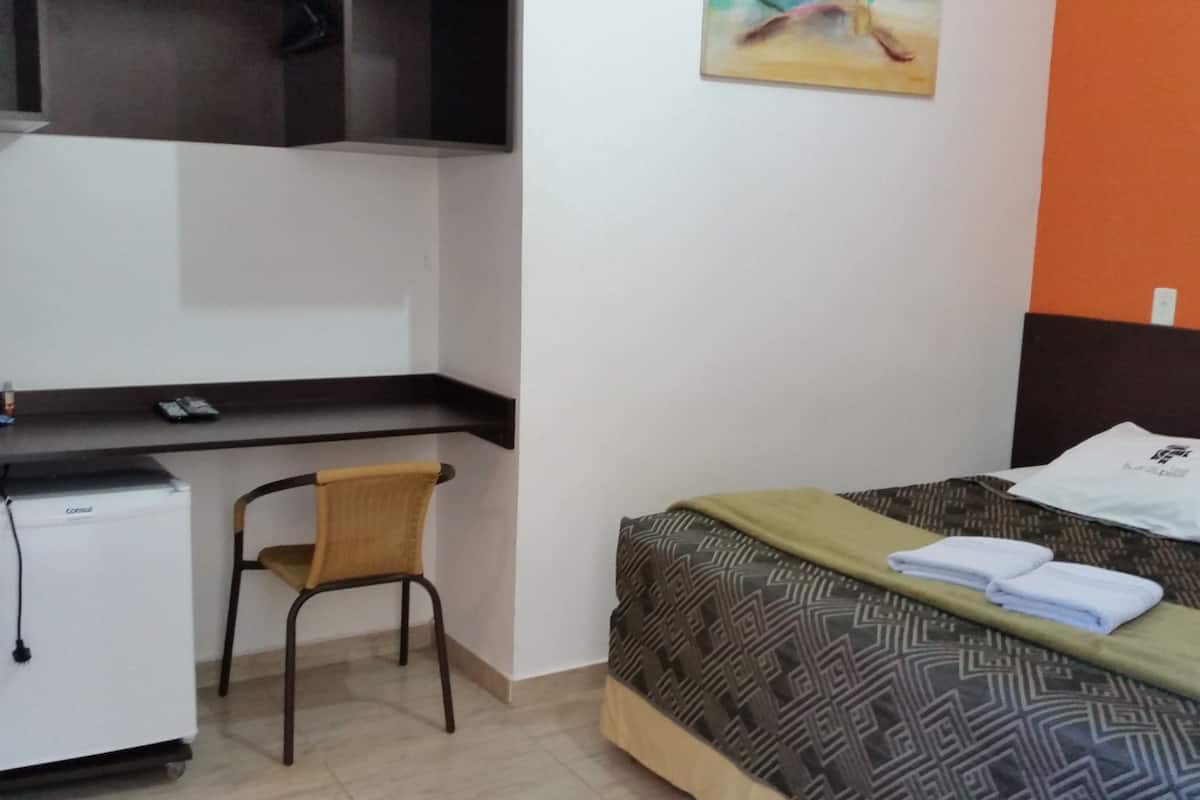A compact hotel room with a bed, a desk with a refrigerator, a chair, and a painting on the wall.