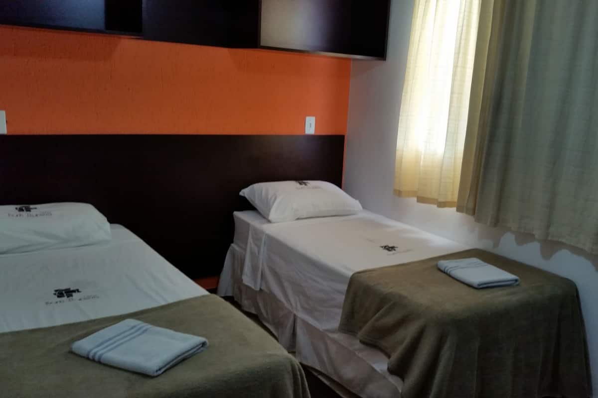 A hotel room with two single beds, each with a white bedspread and a towel. The room has a simple layout with a wooden headboard and a window with curtains.