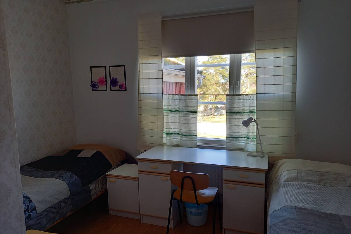 A small, compact room with a single bed, a desk with a chair, and a window with blinds.