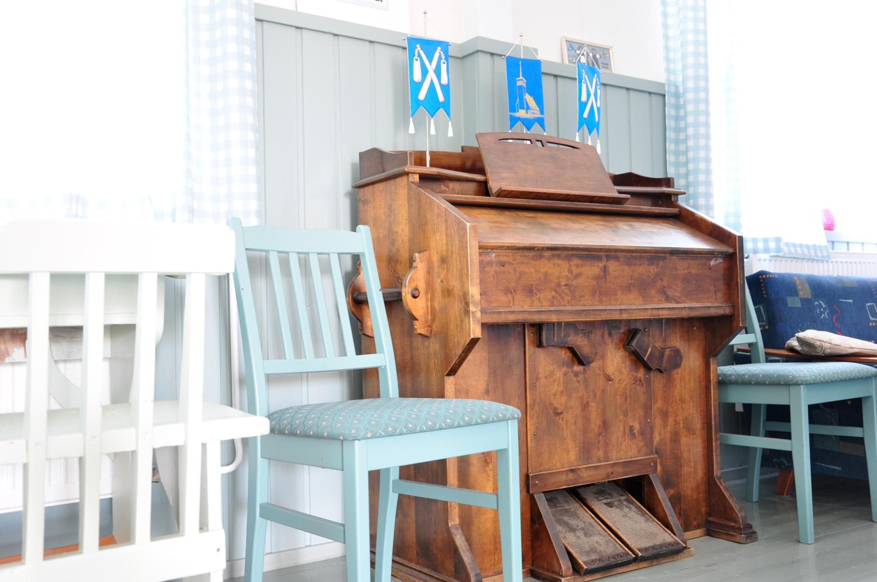 A wooden piano with a bench, a white chair, and a blue chair in a room with a checkered wall.