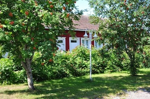 Um edifício vermelho com detalhes em branco, rodeado por vegetação exuberante e árvores carregadas de frutos vermelhos.