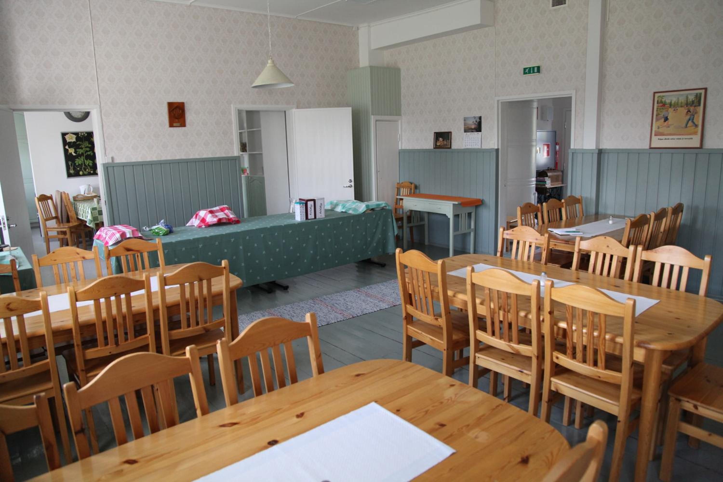 Une pièce avec plusieurs tables et chaises en bois, un lit recouvert d’une couverture verte et une étagère fixée au mur, ornée de livres et d’un tableau encadré.