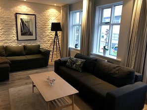 A modern living room with a stone accent wall, a framed picture, a floor lamp, and a view of the street through large windows.