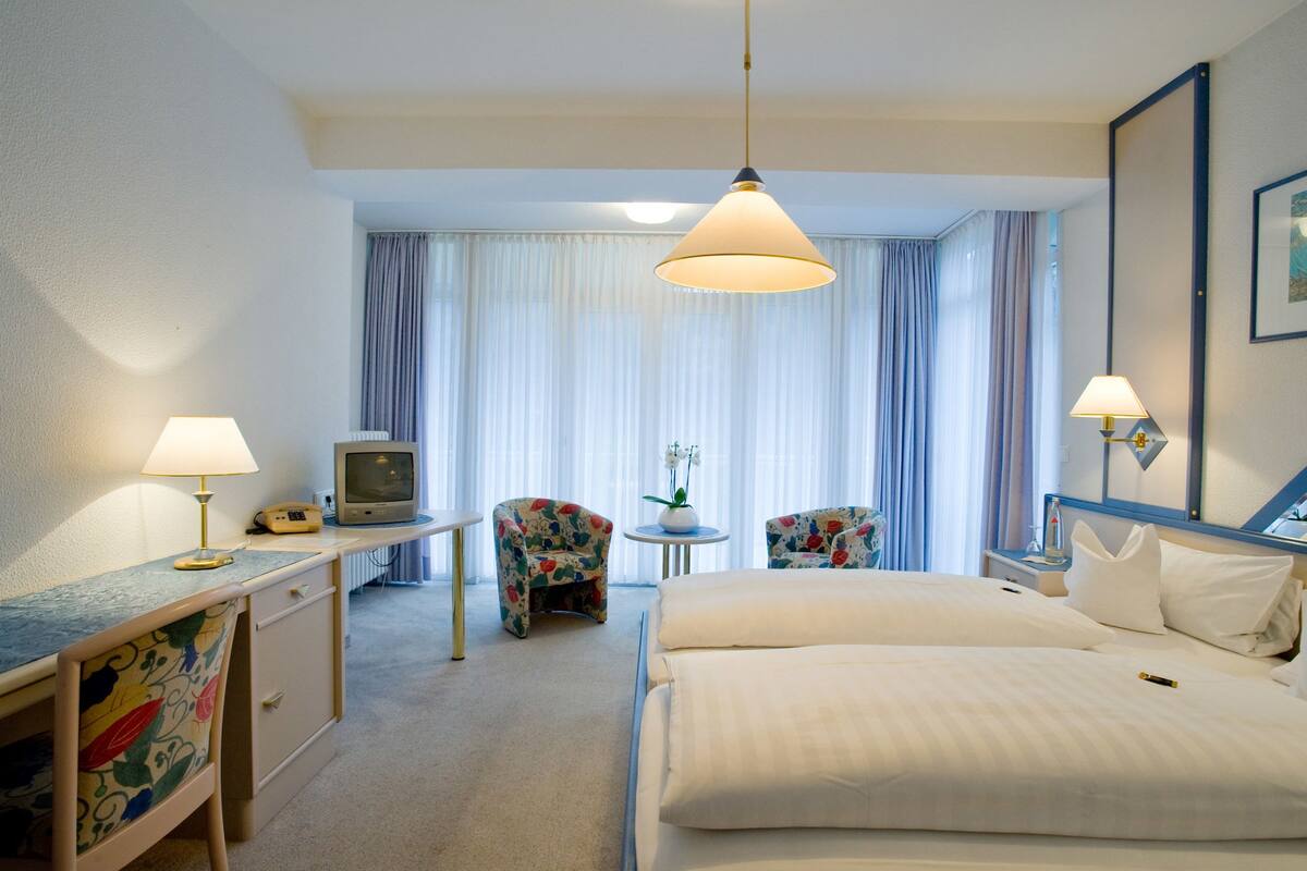 A hotel room with a bed, two chairs, a desk, a television, and a window with curtains.