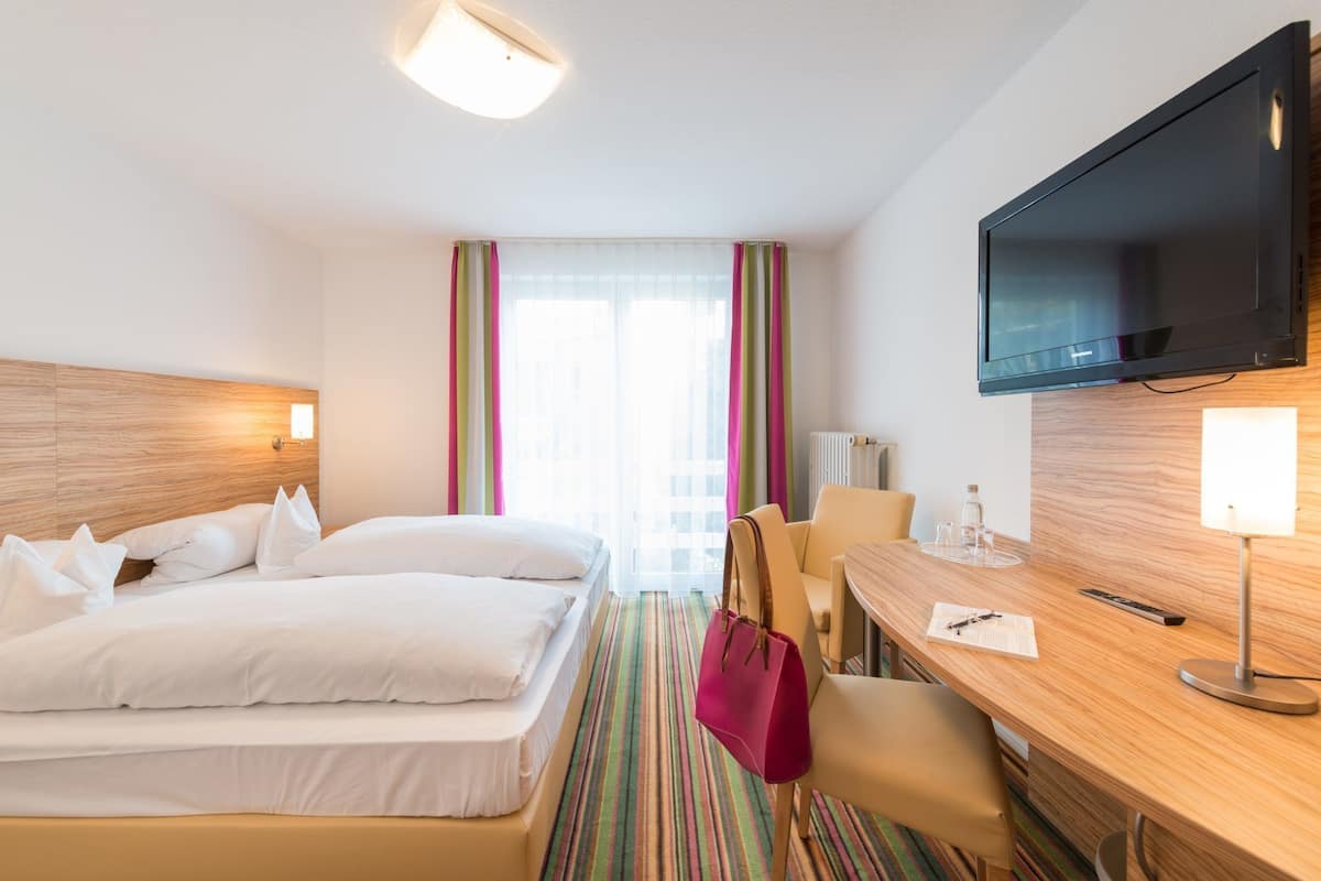 A hotel room with a bed, a desk with a chair, a television, and a window with curtains.