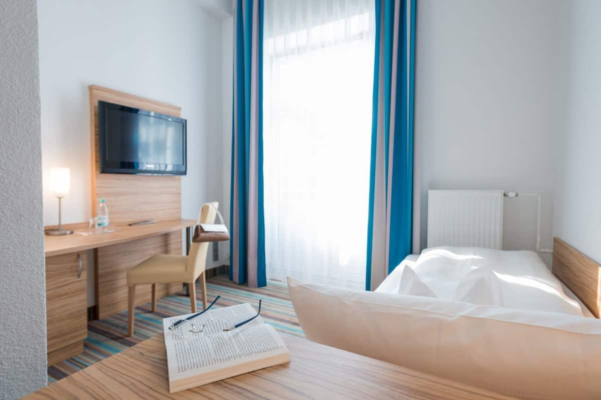 A hotel room with a bed, a desk with a TV, a chair, and a window with curtains.