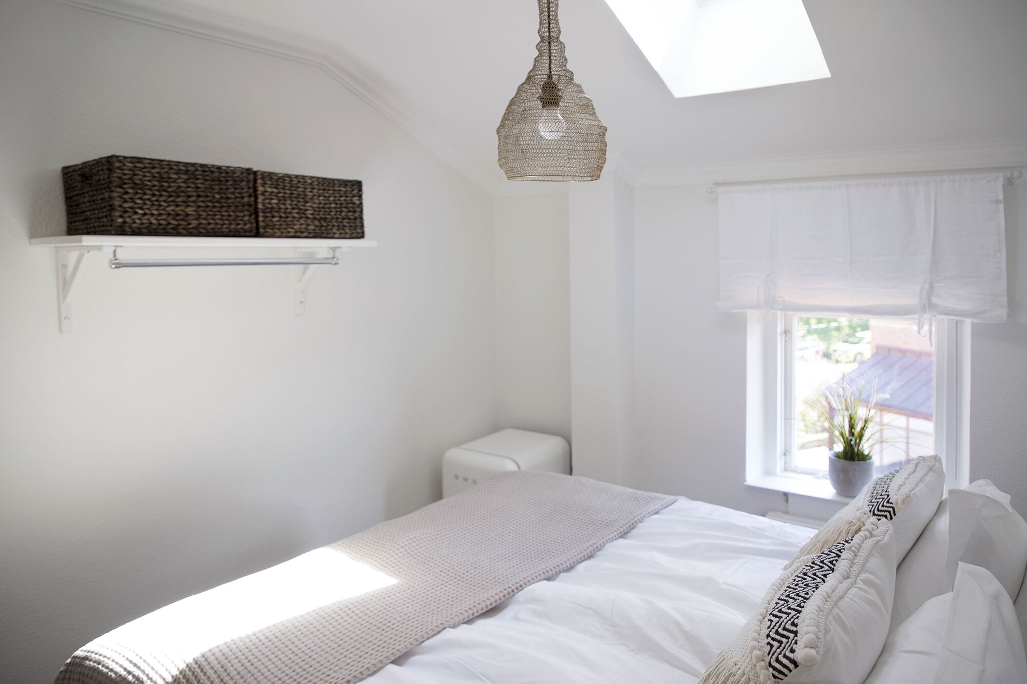 A bedroom with a bed, a hanging light fixture, a window with a view, and a shelf with a basket.