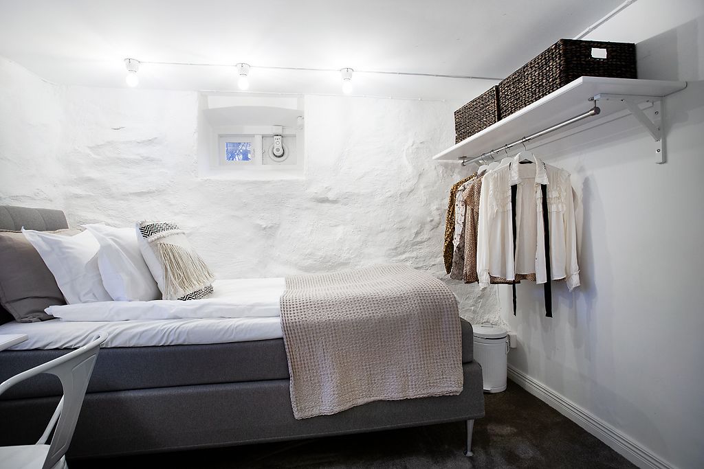 Quarto com cama, mesa de cabeceira, luminária suspensa, janela pequena e estante com roupas.