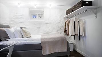 Quarto com cama, mesa de cabeceira, luminária suspensa, janela pequena e estante com roupas.