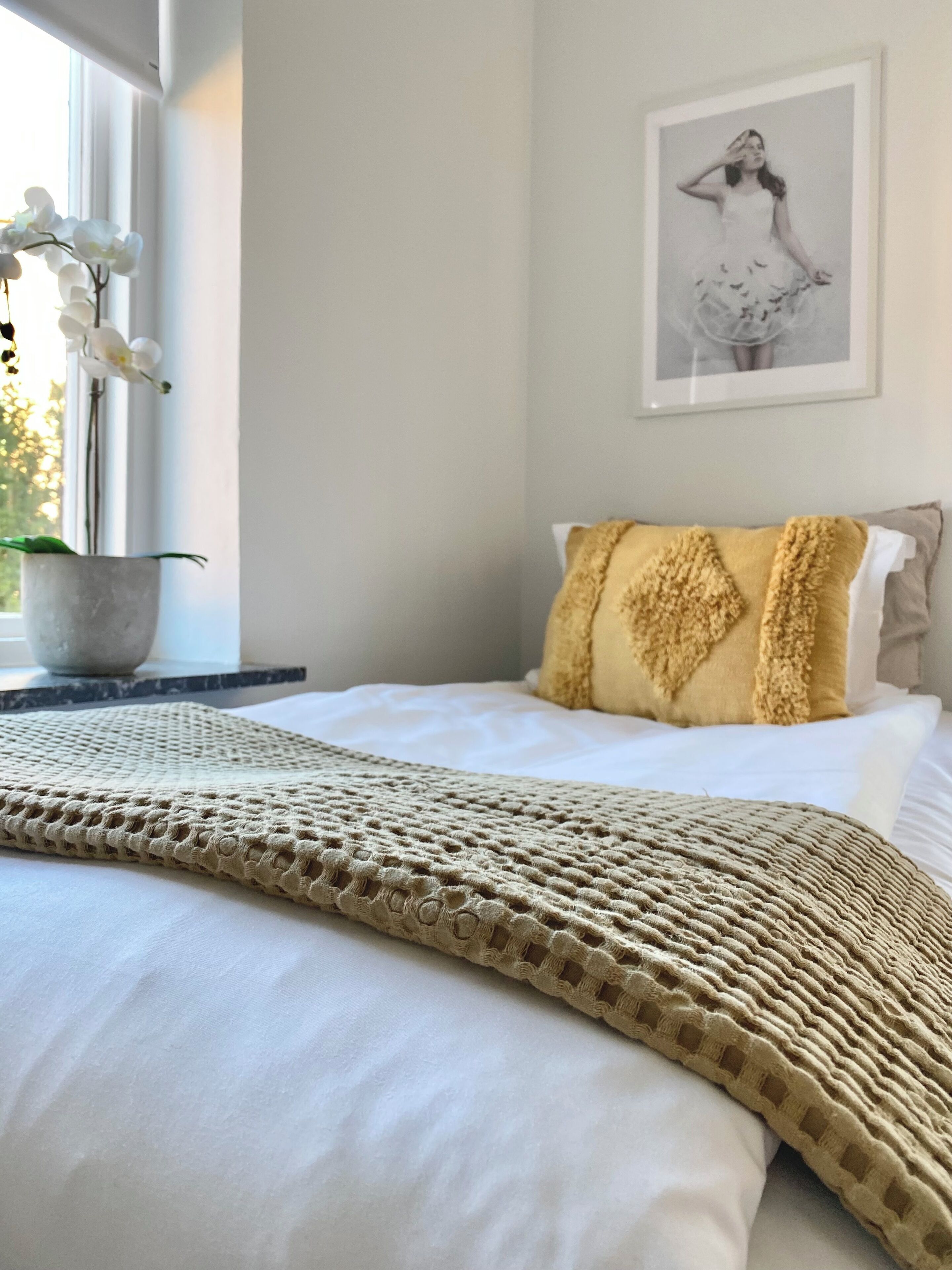 A neatly made bed with a textured blanket, a window with white orchids, and a framed picture on the wall.