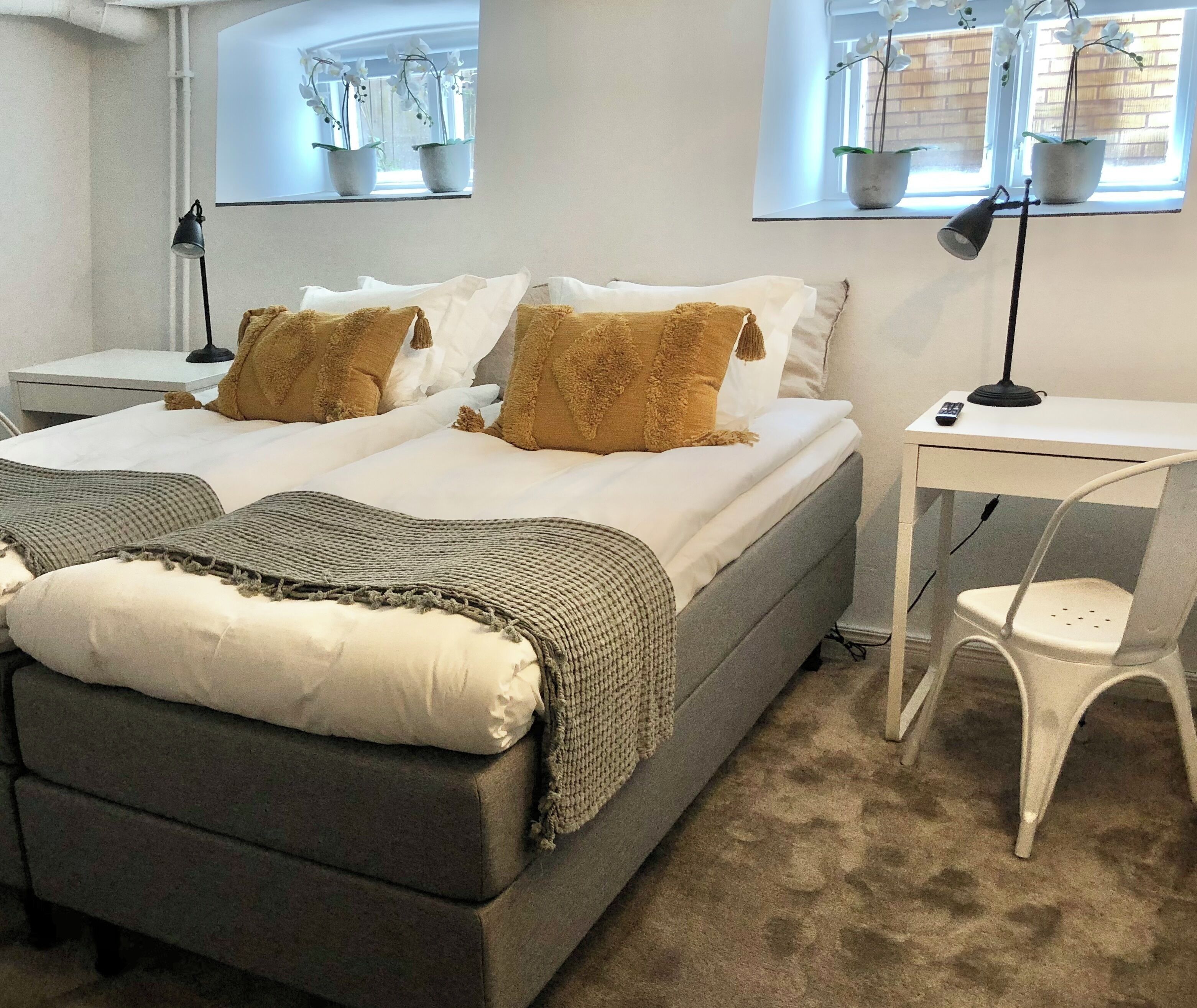 Un dormitorio con cama, mesitas de noche, escritorio, silla y plantas en macetas junto a la ventana.