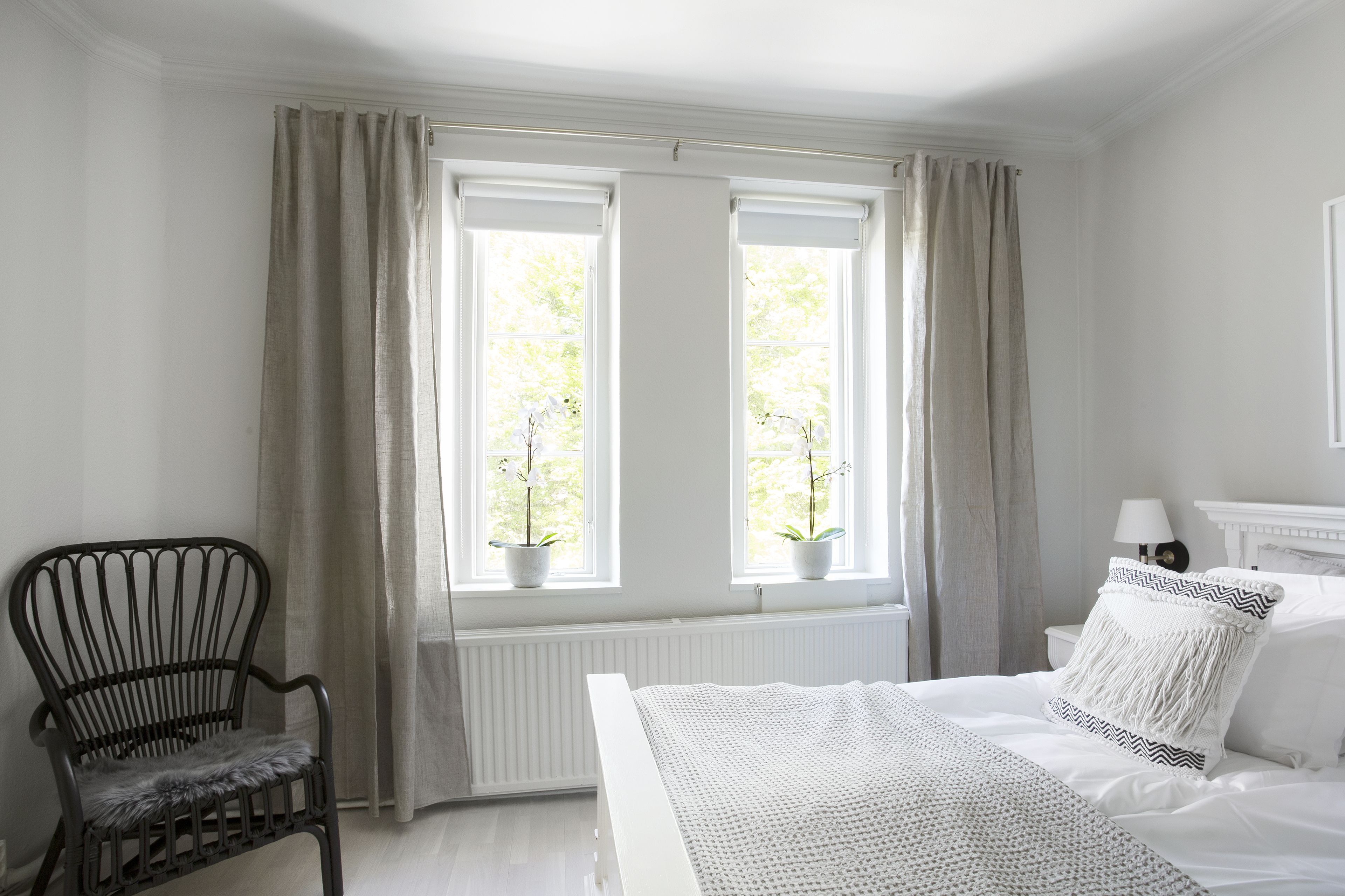 A bedroom with a bed, a chair, two windows with curtains, and a radiator.