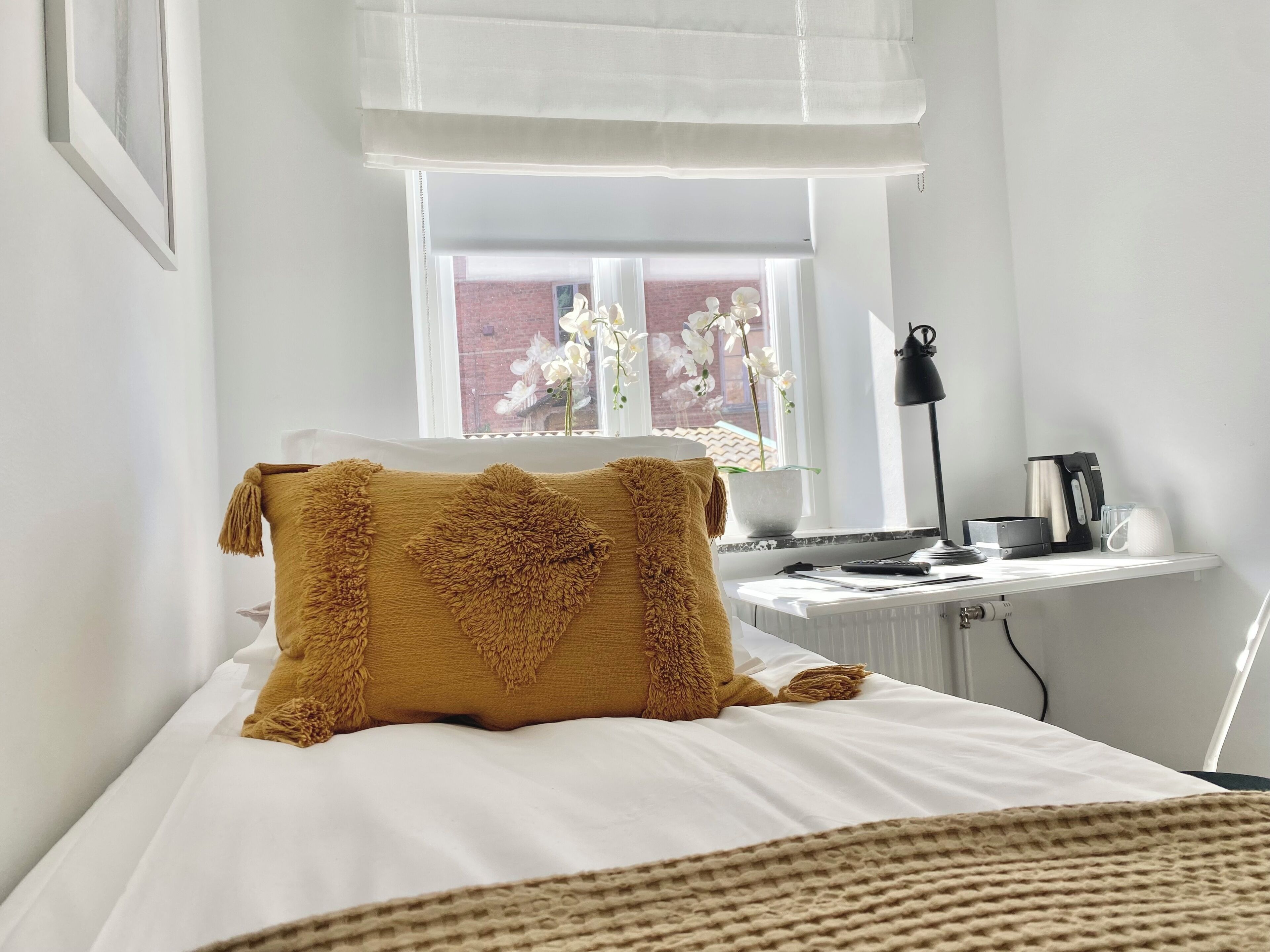 A neatly made bed with a large, textured pillow, a bedside table with a lamp, a coffee maker, and a view of a brick wall through a window with white curtains.