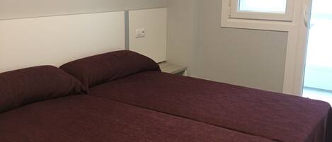 A room with two single beds, a small bedside table, a nightstand, a window with blinds, and a wall-mounted artwork.