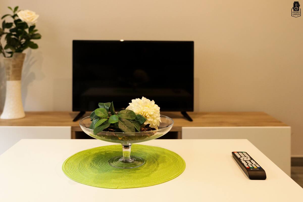 A modern living room with a white table, a glass bowl with flowers, a remote control, and a TV on a stand.