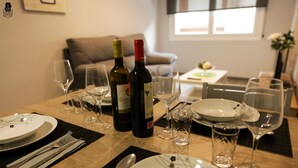 Una mesa de comedor dispuesta con platos, cubiertos, botellas de vino y vasos, en una habitación bien iluminada con un sofá y una ventana al fondo.