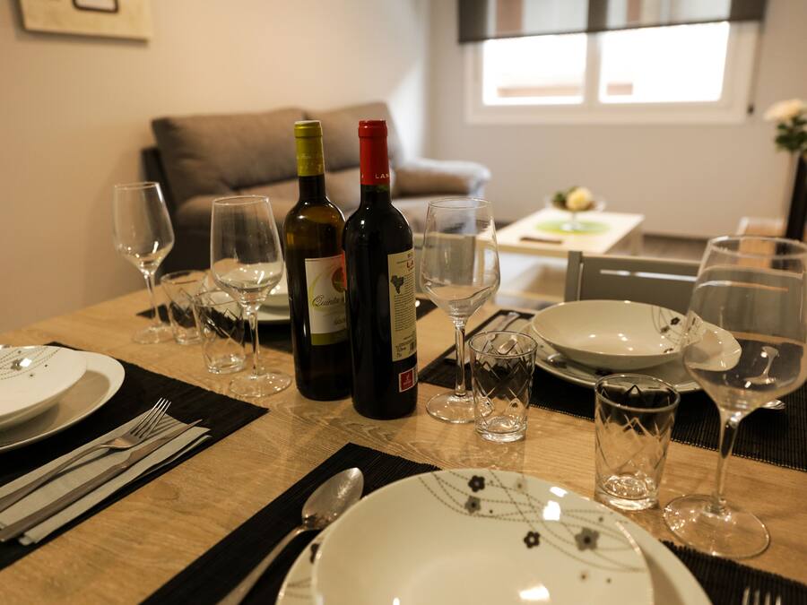 Una mesa de comedor dispuesta con platos, cubiertos, botellas de vino y vasos, en una habitación bien iluminada con un sofá y una ventana al fondo.
