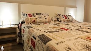 Una cama con colcha que muestra sellos y mapas diversos, una mesita de noche con lámpara, y una ventana con cortinas.