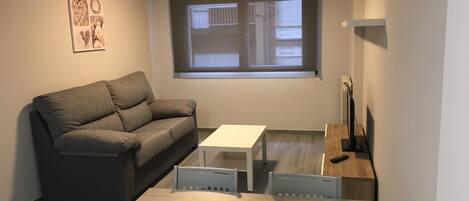 Un espacio habitable pequeño y moderno con un sofá gris, una mesa blanca y un televisor pequeño.