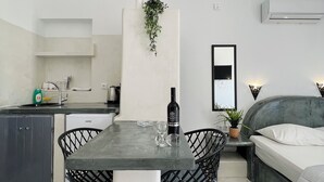 A modern kitchen with a dining table, chairs, and a hanging plant. There is a bed with a headboard and a bedside table with a plant. - Santo Margarita (Santorini)