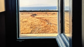 A window with a view of a dry, grassy field and distant mountains. - Santo Margarita (Santorini)