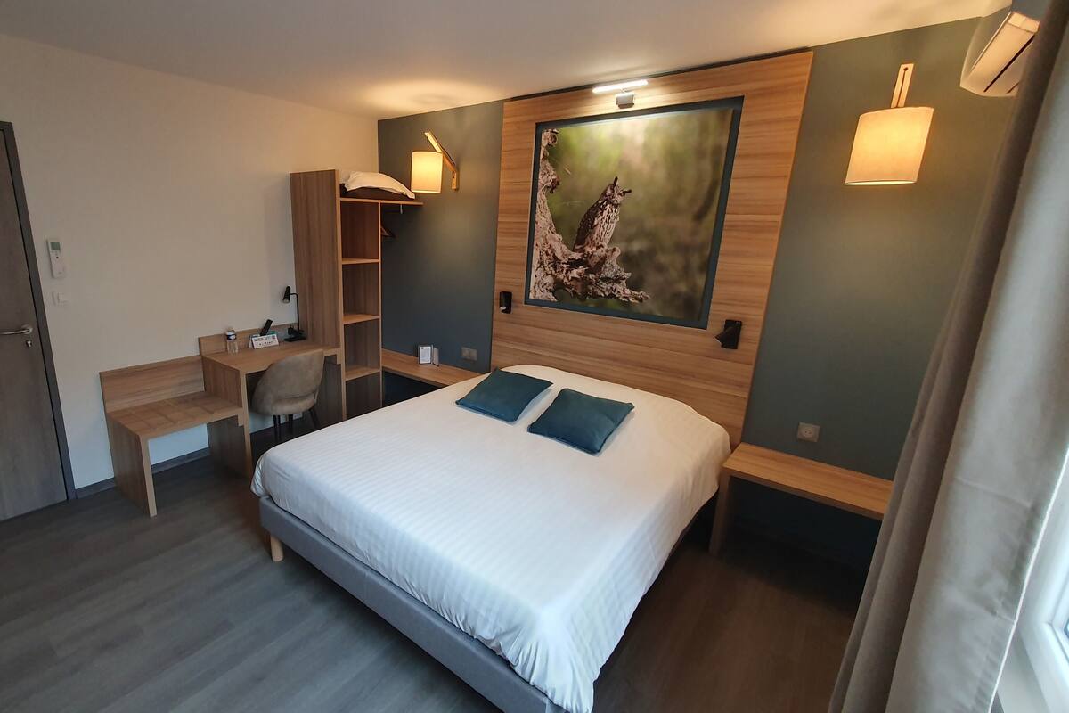 A hotel room with a bed, desk, chair, and a painting on the wall.