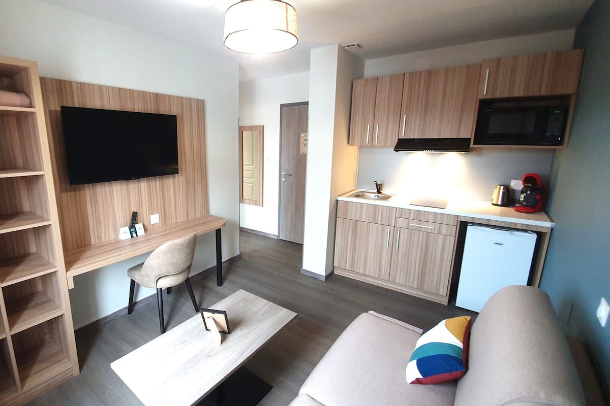 A compact living space with a kitchenette, a flat-screen TV mounted on the wall, a sofa with a colorful cushion, and a wooden shelving unit.