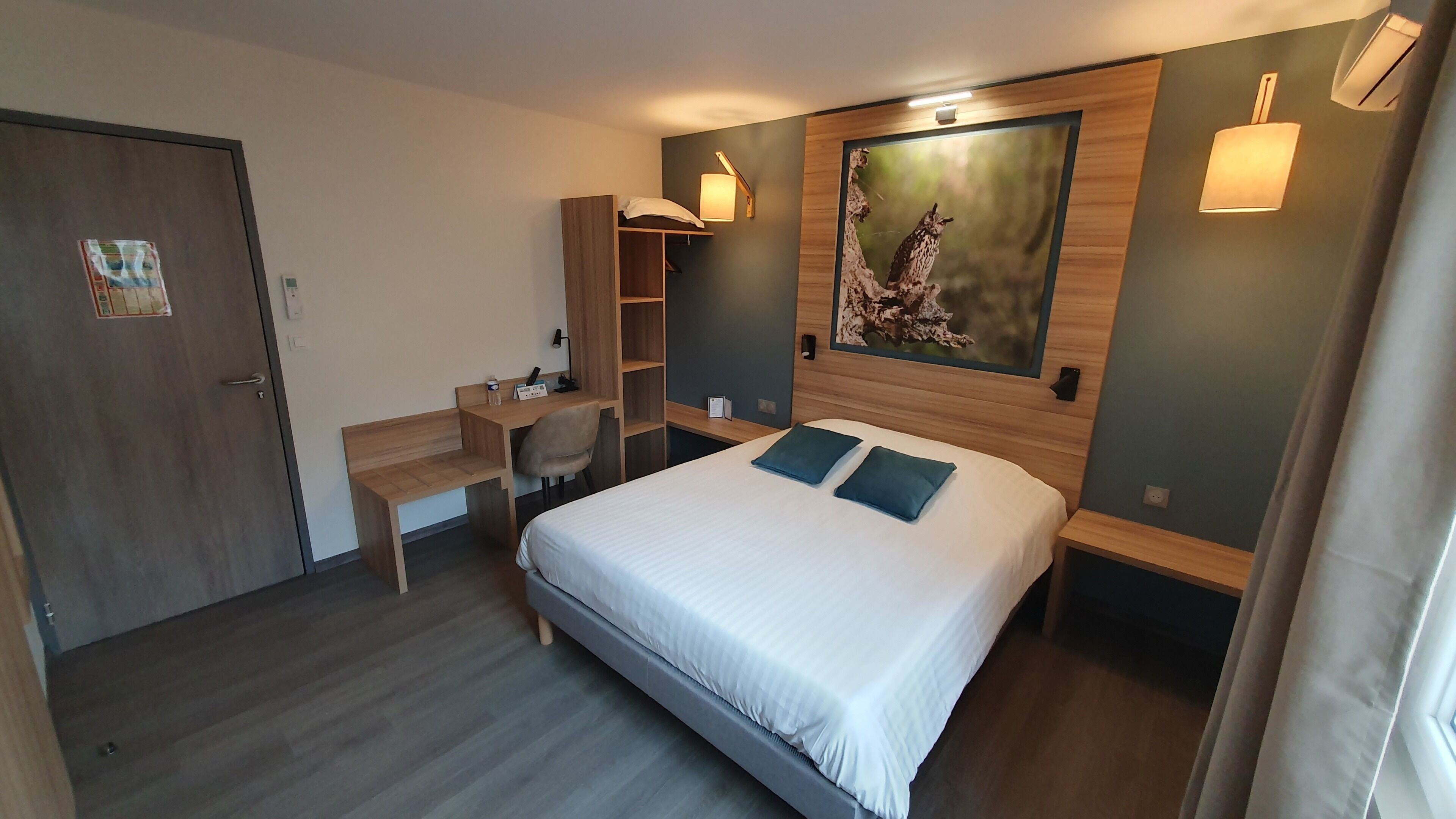 A hotel room with a bed, desk, chair, and a painting on the wall.