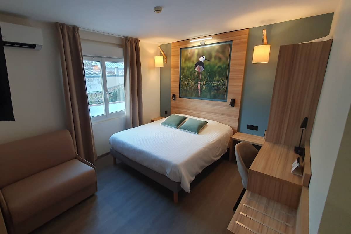 A hotel room with a bed, a seating area, a desk, and a painting on the wall.