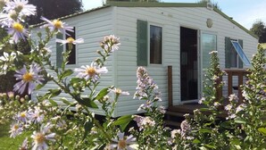 A mobile home with a deck and a covered porch, surrounded by blooming flowers. - Camping des Platanes (Charny-Oree-de-Puisaye)