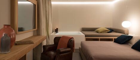 A modern hotel room with a wooden vanity, a leather armchair, a sofa, a bed, and a large mirror.