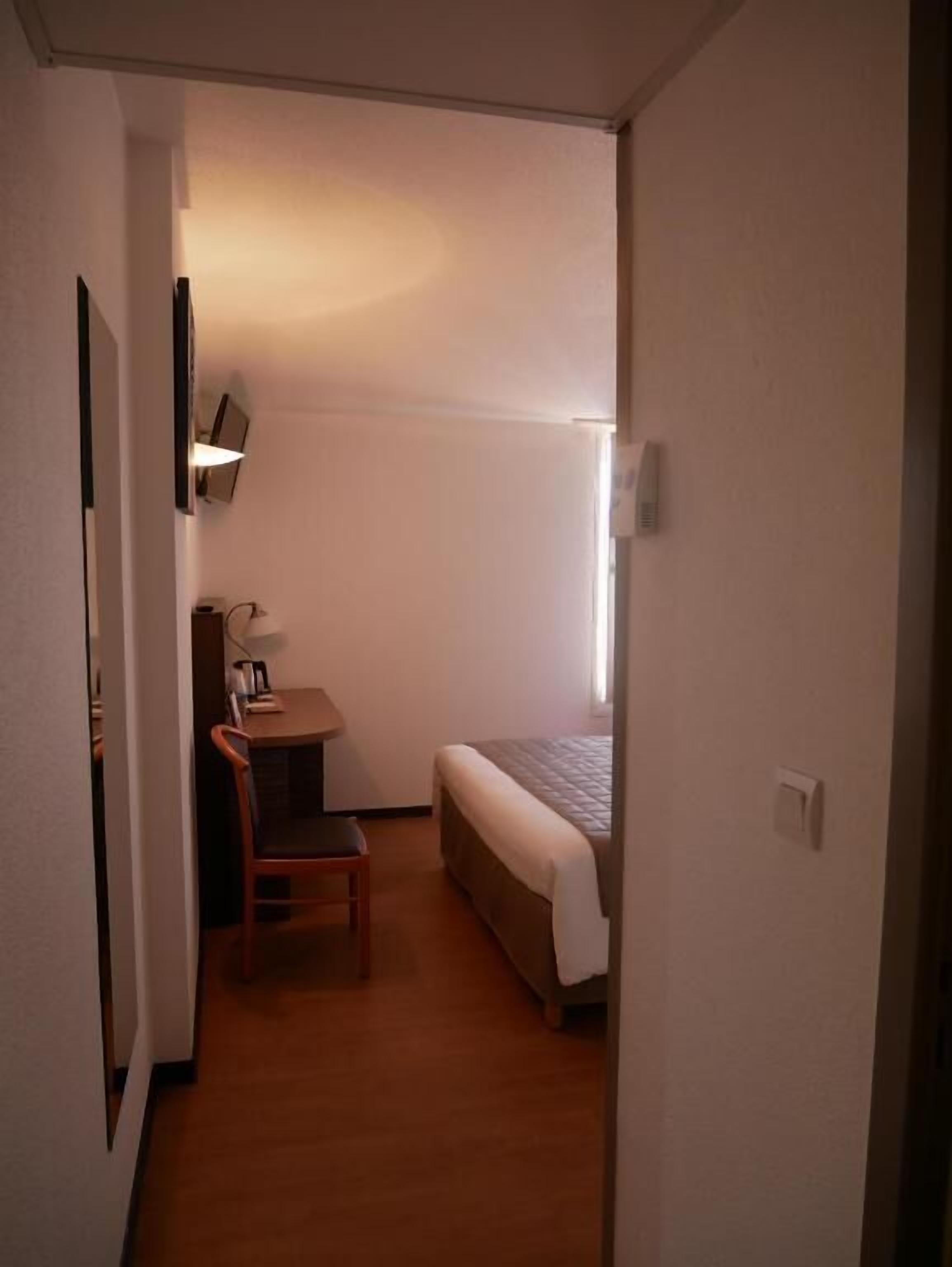 A small, compact room with a bed, a desk, and a chair. There is a window providing natural light, and a wall-mounted light fixture.