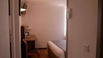 A small, compact room with a bed, a desk, and a chair. There is a window providing natural light, and a wall-mounted light fixture.