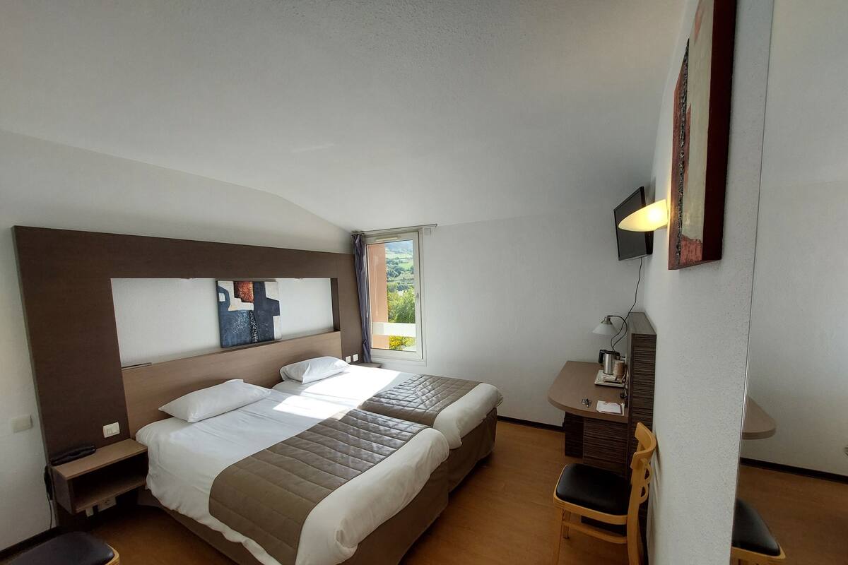 Habitación de hotel con dos camas, un escritorio, una silla, una mesita de noche y ventana con vista a zonas verdes.