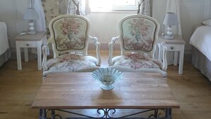 Une pièce avec un mur en pierre, deux fauteuils recouverts d’un tissu à motifs floraux, une table en bois sur laquelle repose un saladier en verre, et un lit surmonté d’un baldaquin blanc.