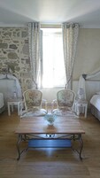 Una habitación con pared de piedra, dos sillones tapizados con motivos florales, una mesa de madera con un cuenco de vidrio y una cama con dosel blanco.